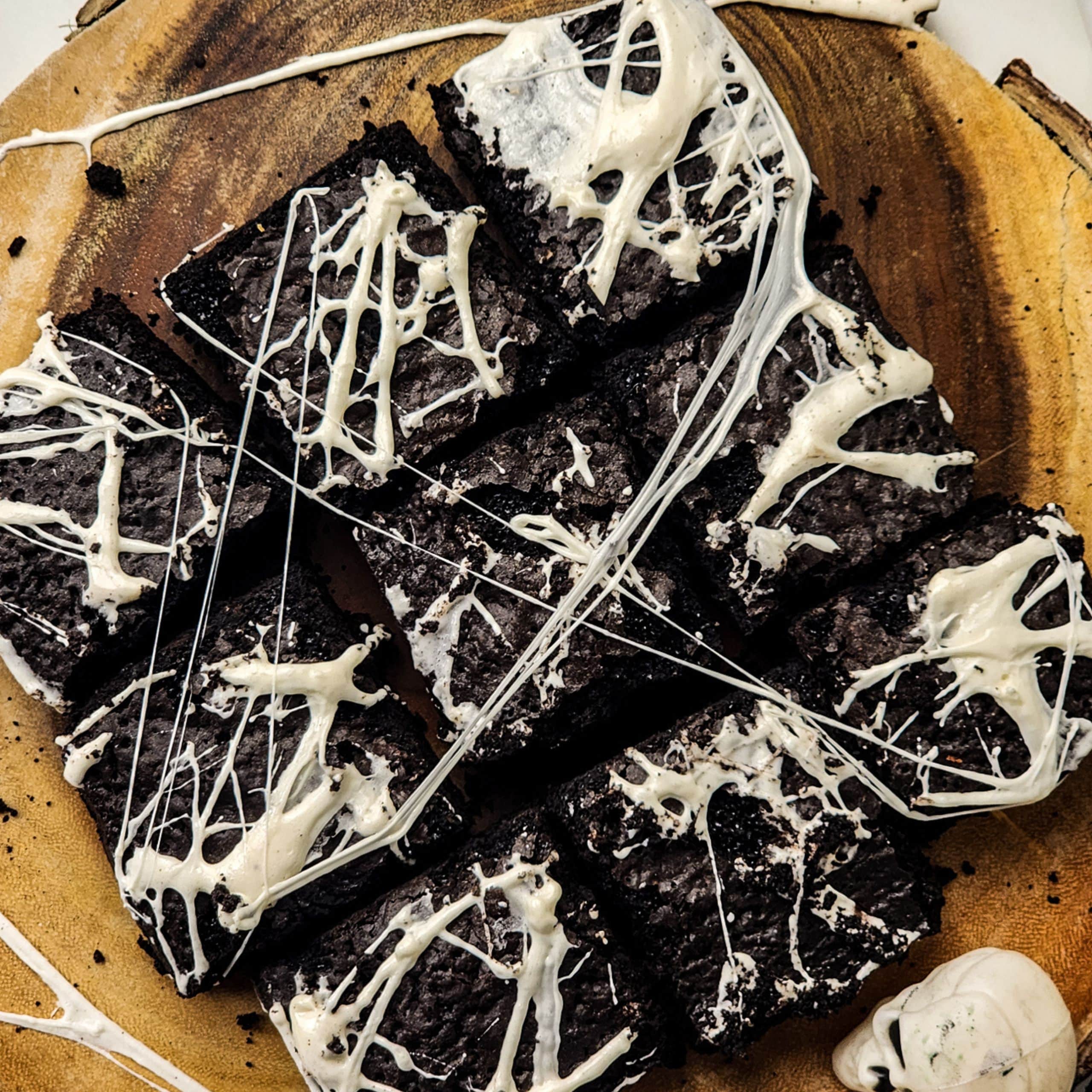 A wooden plate with nine Spiderweb Brownies topped with drizzled white icing, arranged closely together.