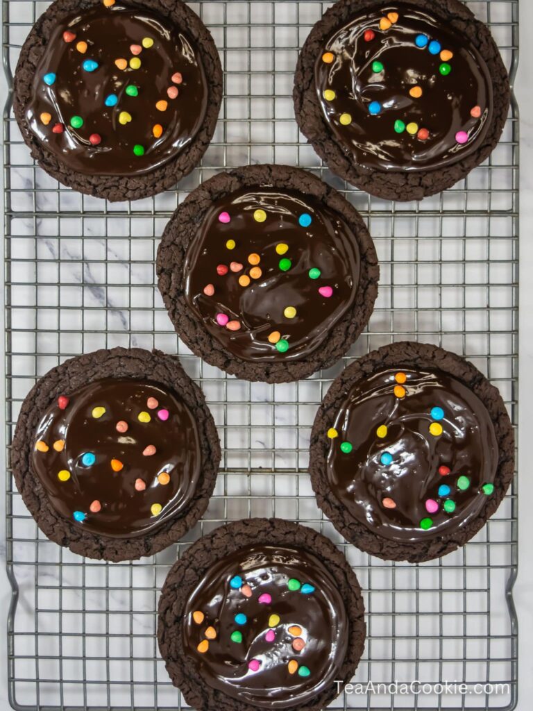 A wire rack with chocolate cosmic brownie cookies topped with chocolate ganache and colorful candies.