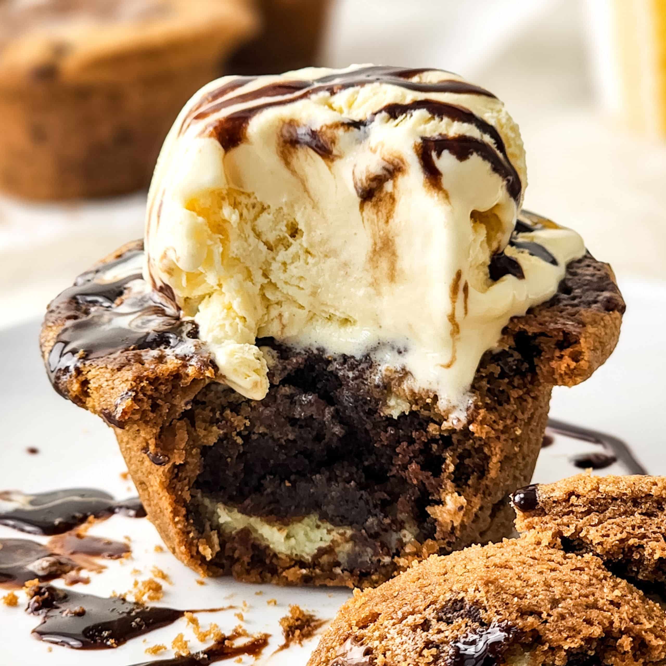 A chocolate chip cookie cup filled with brownie, topped with a scoop of vanilla ice cream and drizzled with chocolate syrup on a white plate.