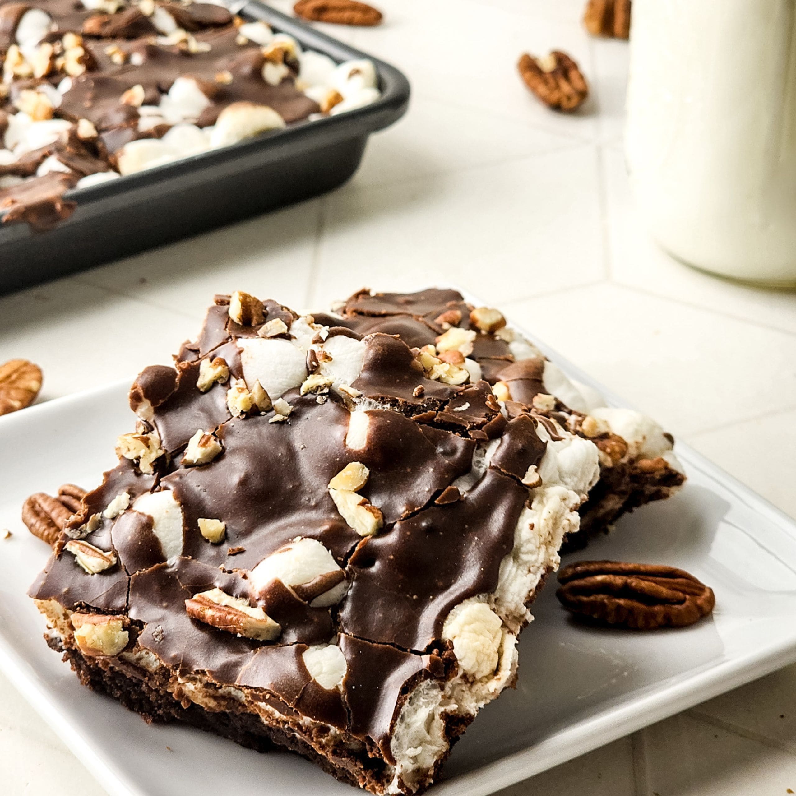 Two squares of chocolate brownie topped with marshmallows and chopped pecans on a white plate, with a baking tray and a glass of milk in the background.