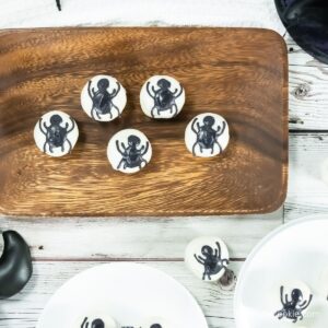 Five white treats with black spider decorations are arranged on a wooden tray, with additional treats on white plates nearby, set on a white wooden surface.