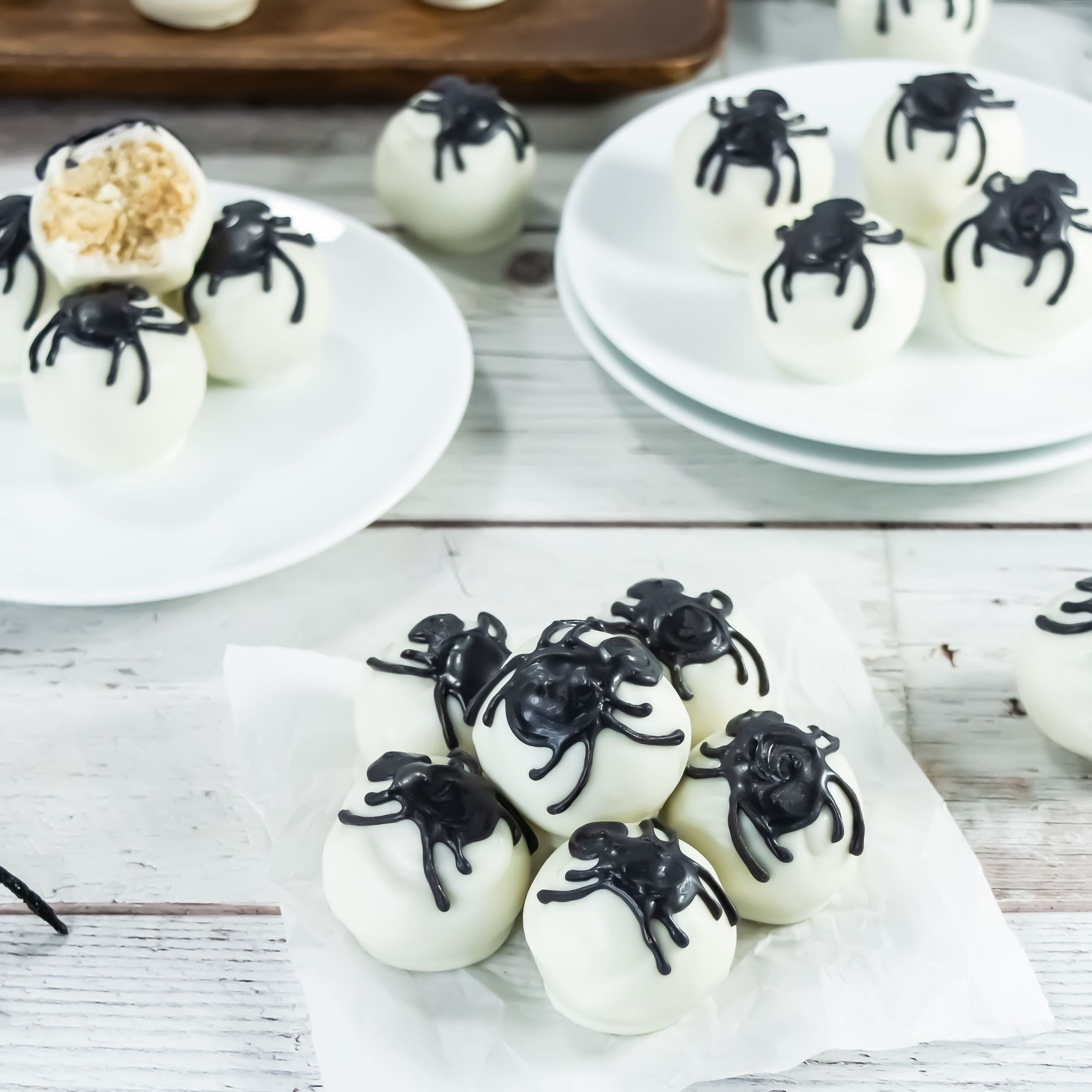 White chocolate truffles decorated with black spider designs are arranged on plates and parchment paper on a white wooden surface.