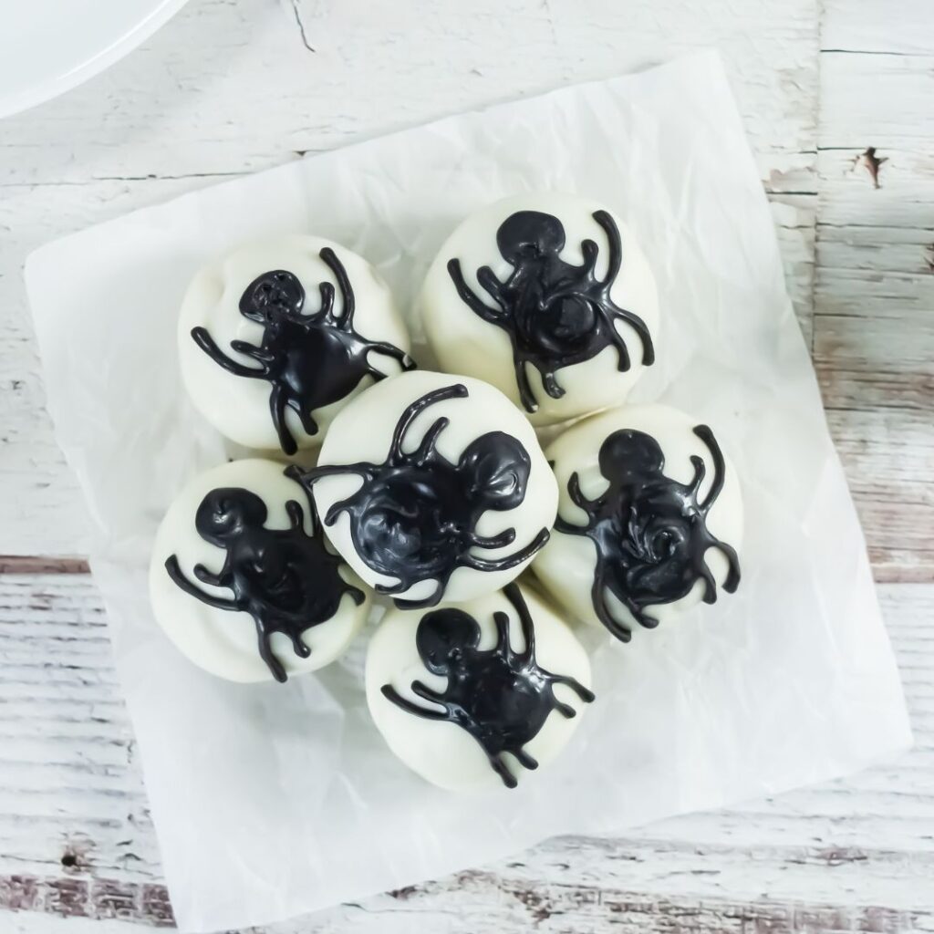 Cake ball spiders. Six white round treats topped with black spider designs made of icing, arranged on white parchment paper on a rustic wooden surface.