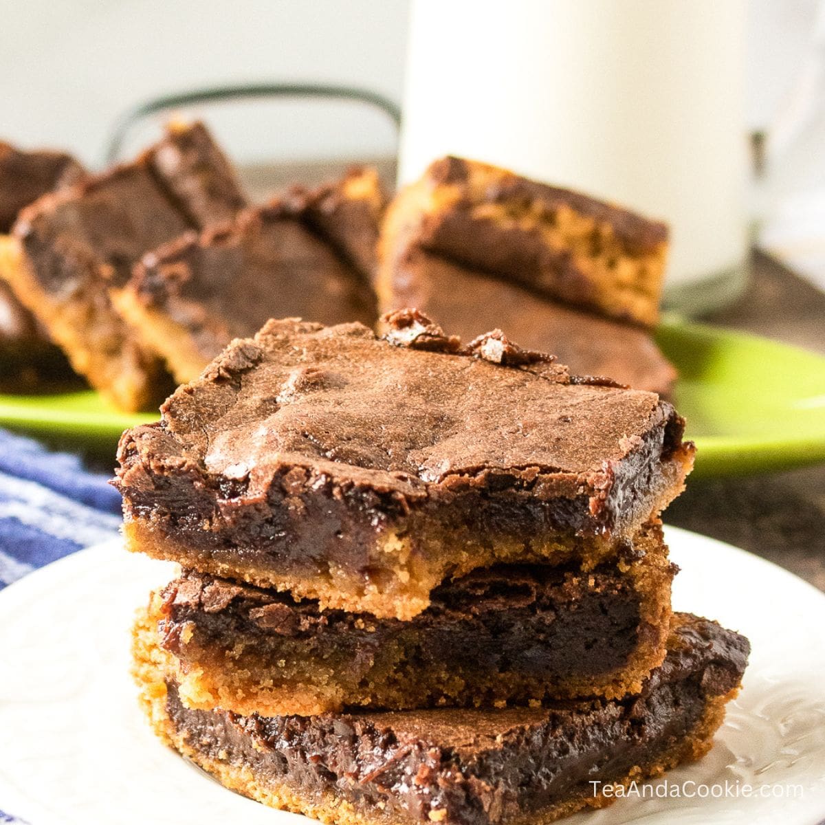 Chocolate Peanut Butter Brownies. Three thick chocolate chip cookie brownie bars stacked on a white plate, with more bars and a glass of milk in the background.