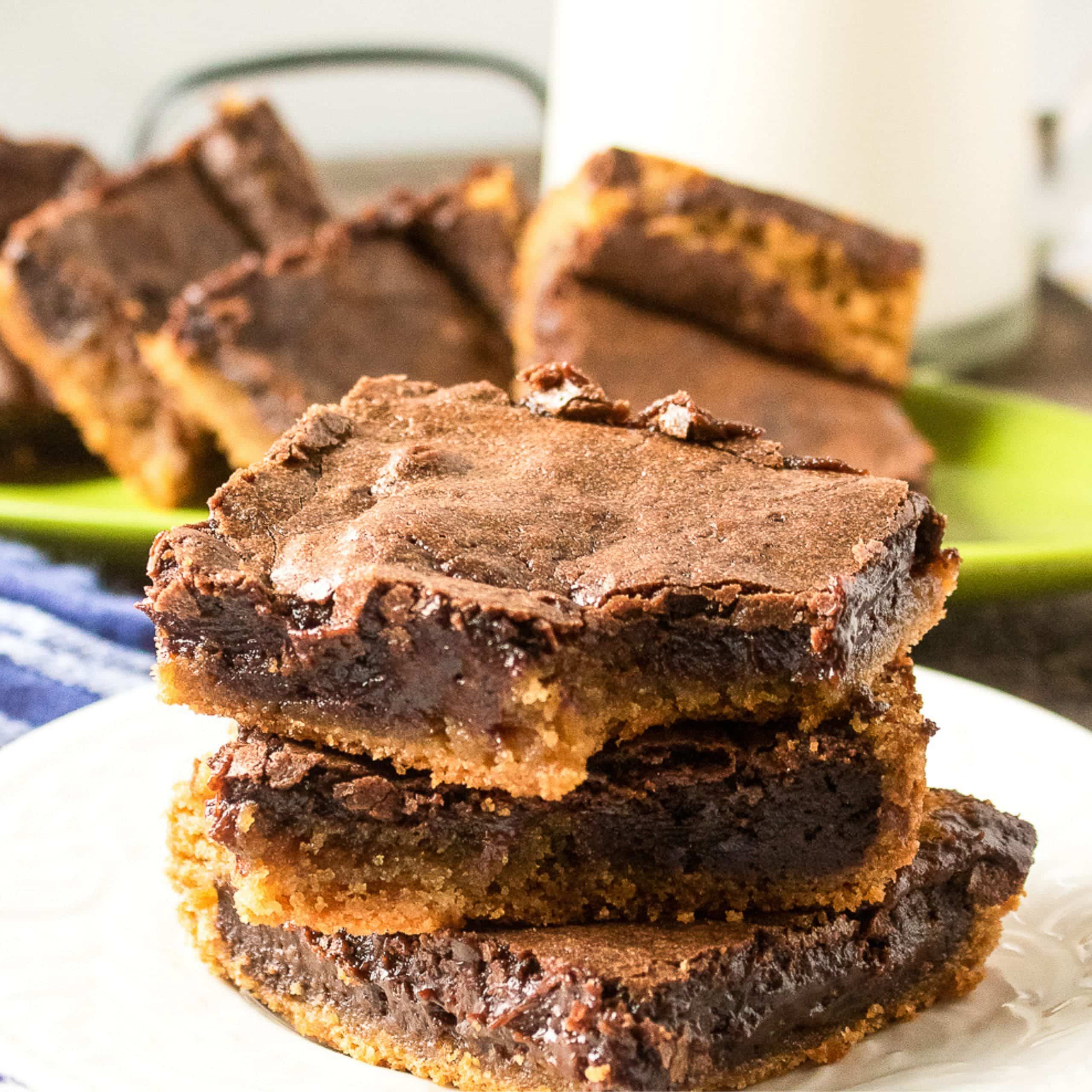 Three chocolate brownies stacked on a white plate, with more brownies on a green plate in the background and a glass of milk.