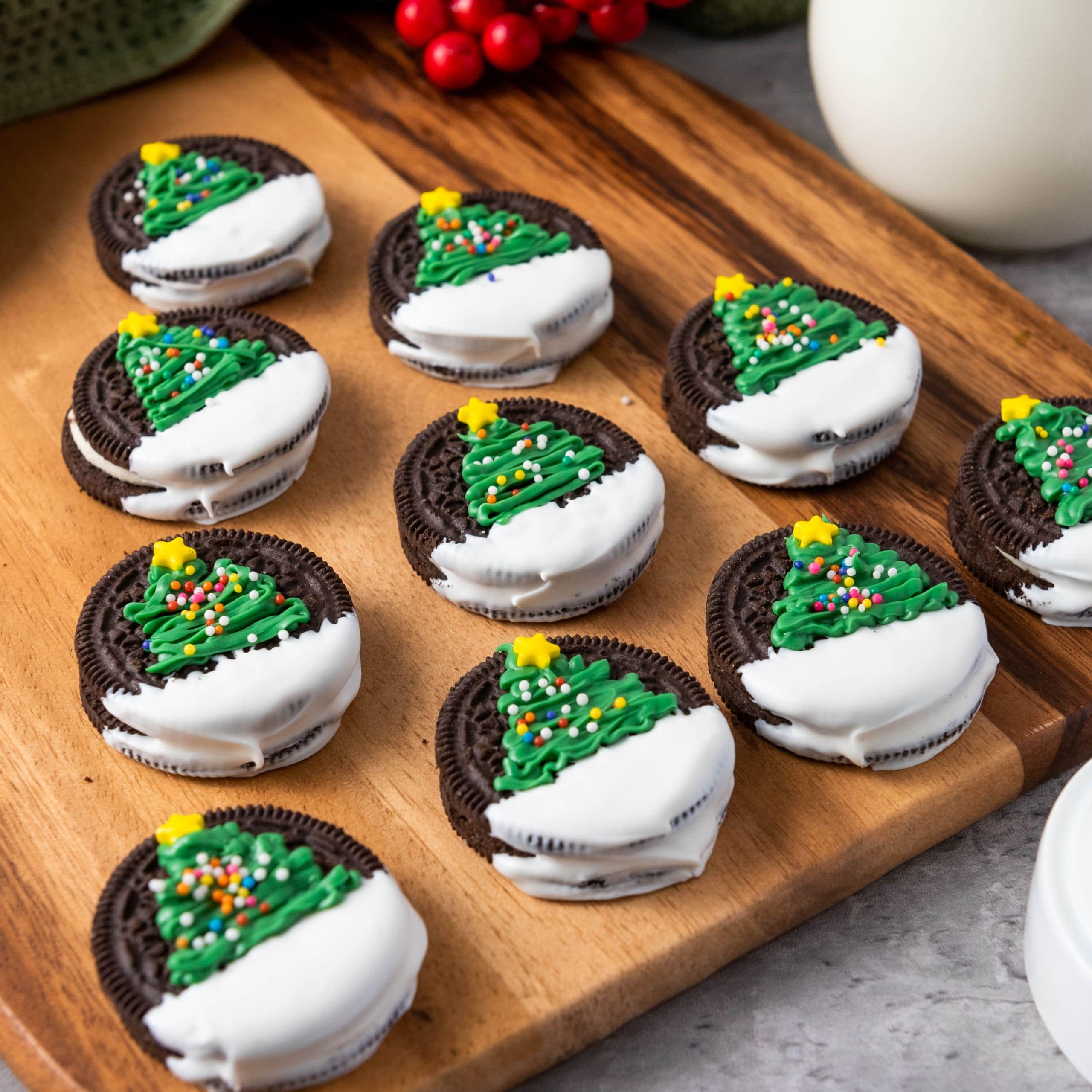 Making Christmas Tree Oreos. Chocolate sandwich cookies half-dipped in white icing, each topped with green icing shaped like a Christmas tree, sprinkles, and a yellow star, arranged on a wooden board.