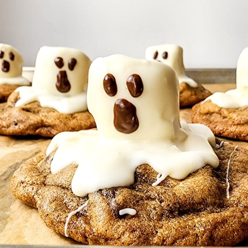 Chocolate chip cookies topped with marshmallows coated in white icing, decorated with chocolate to resemble ghosts.