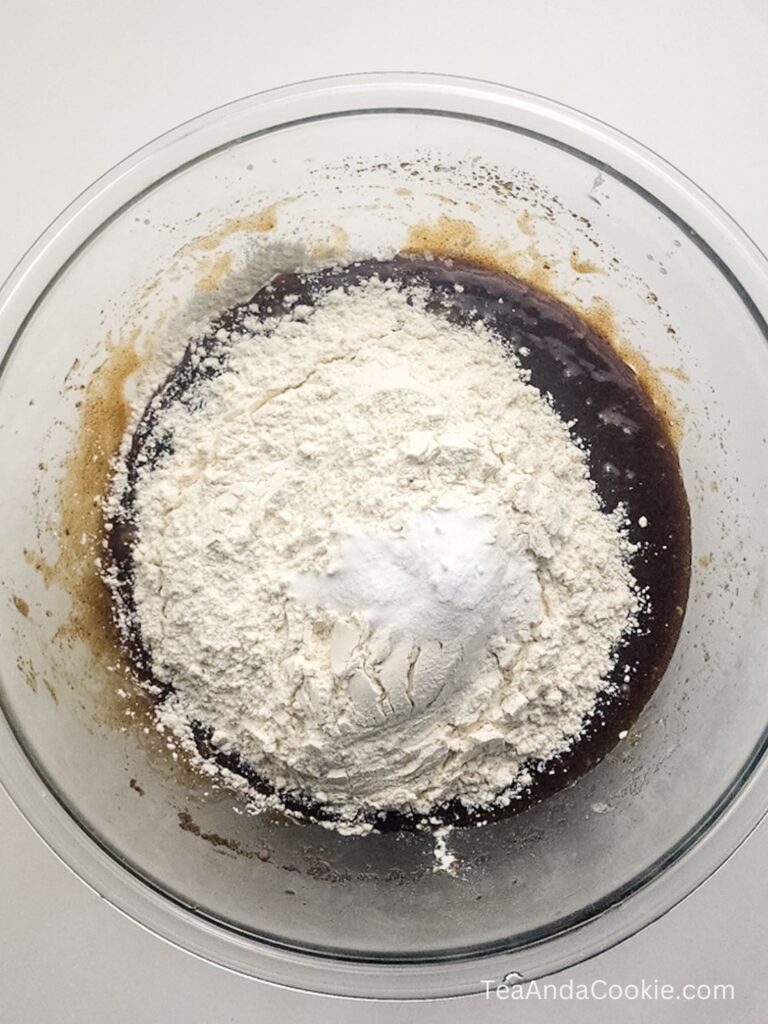 A glass bowl containing brown batter with a mound of white flour and baking powder on top, ready to be mixed.