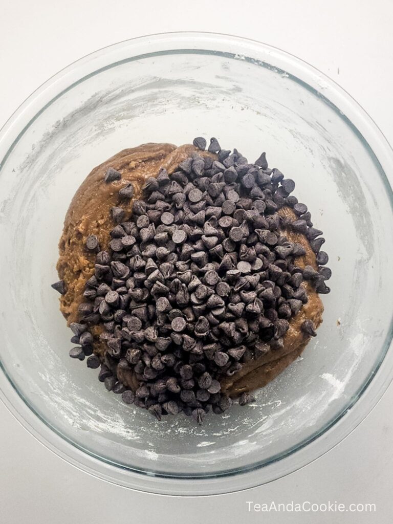 A glass bowl with cookie dough topped with a pile of chocolate chips, ready to be mixed.