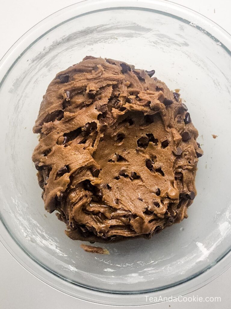 A glass bowl containing a mound of chocolate chip cookie dough on a white surface.