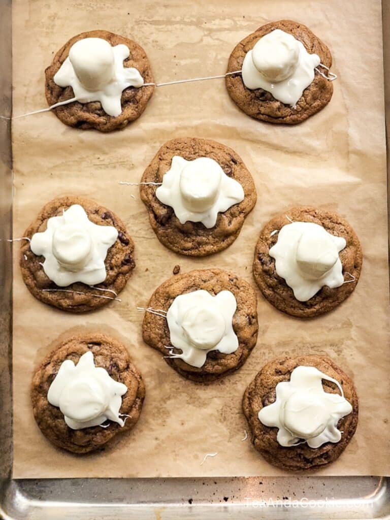 Eight chocolate chip cookies on parchment paper, each topped with a partially melted marshmallow, on a metal baking tray.