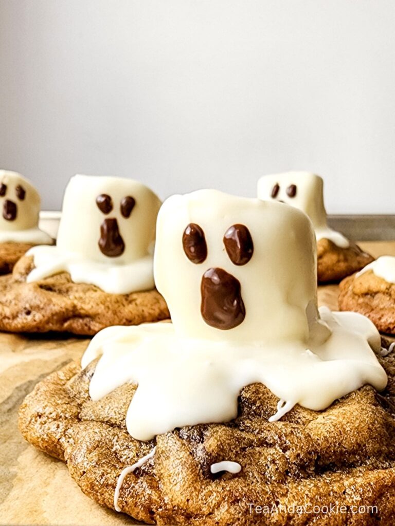 Cookies topped with melted white chocolate and marshmallows decorated with chocolate eyes and mouths to resemble ghosts, placed on parchment paper.