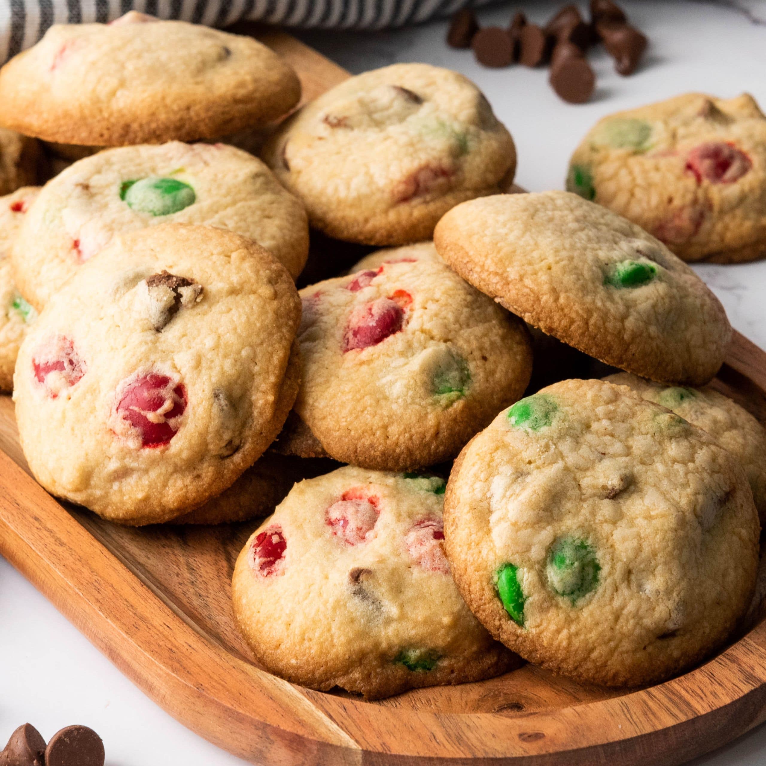 A wooden tray filled with cookies containing red and green candy pieces, with some chocolate chips visible in the cookies and scattered on the surface.