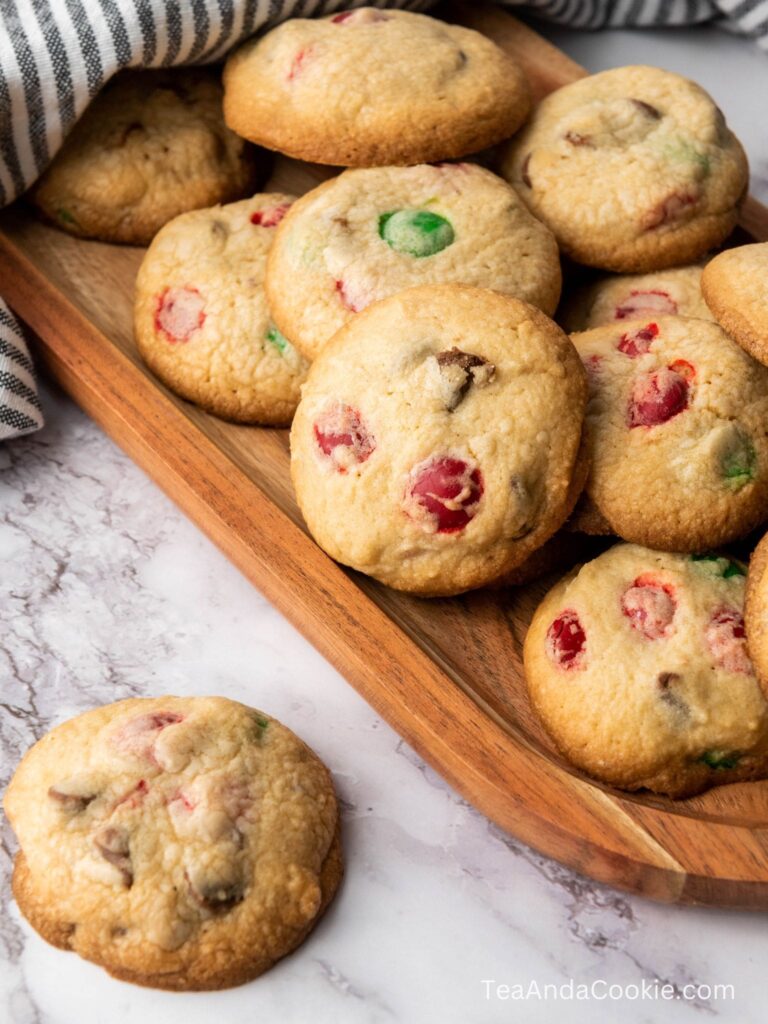 A wooden tray filled with cookies containing red and green candied fruit and chocolate chips, with one cookie placed on a marble surface.