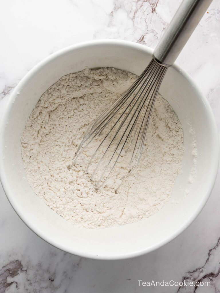 A metal whisk rests in a white bowl filled with flour on a marble surface. The website TeaAndaCookie.com is visible in the corner.