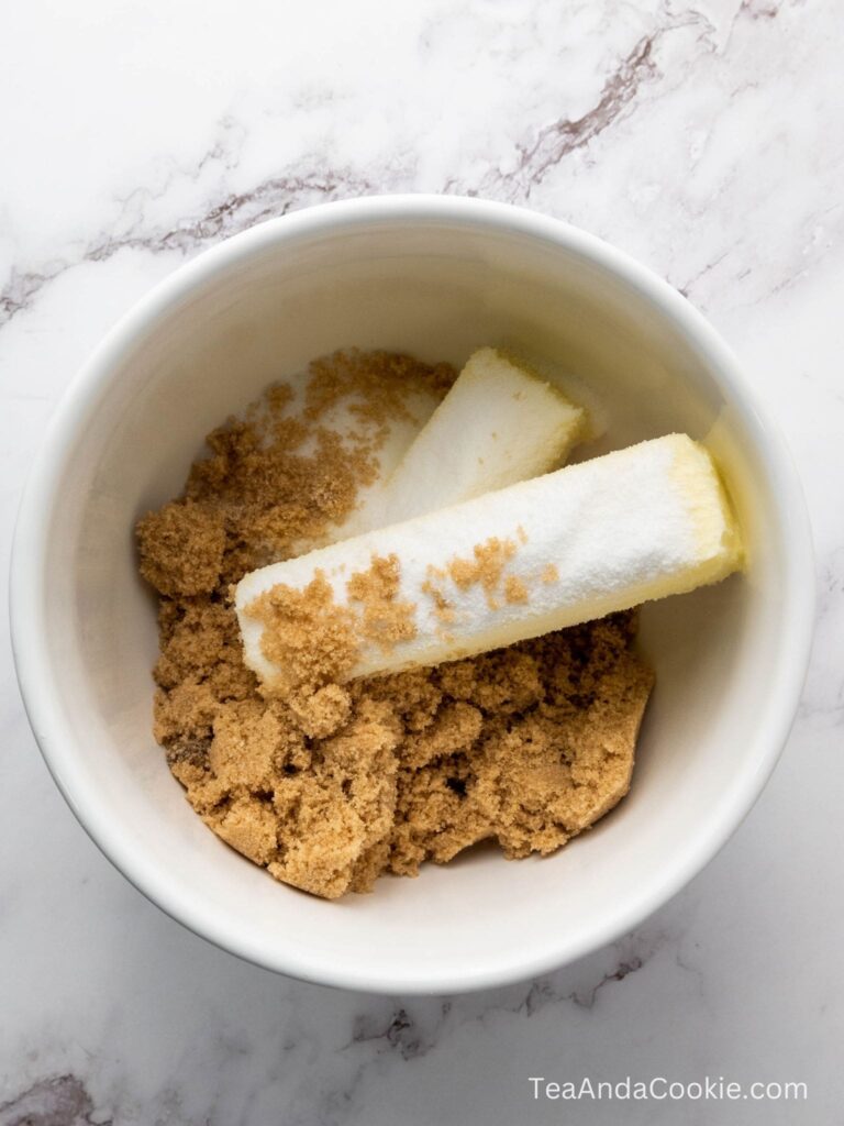 A white bowl contains two sticks of butter, brown sugar, and granulated white sugar on a marble surface.