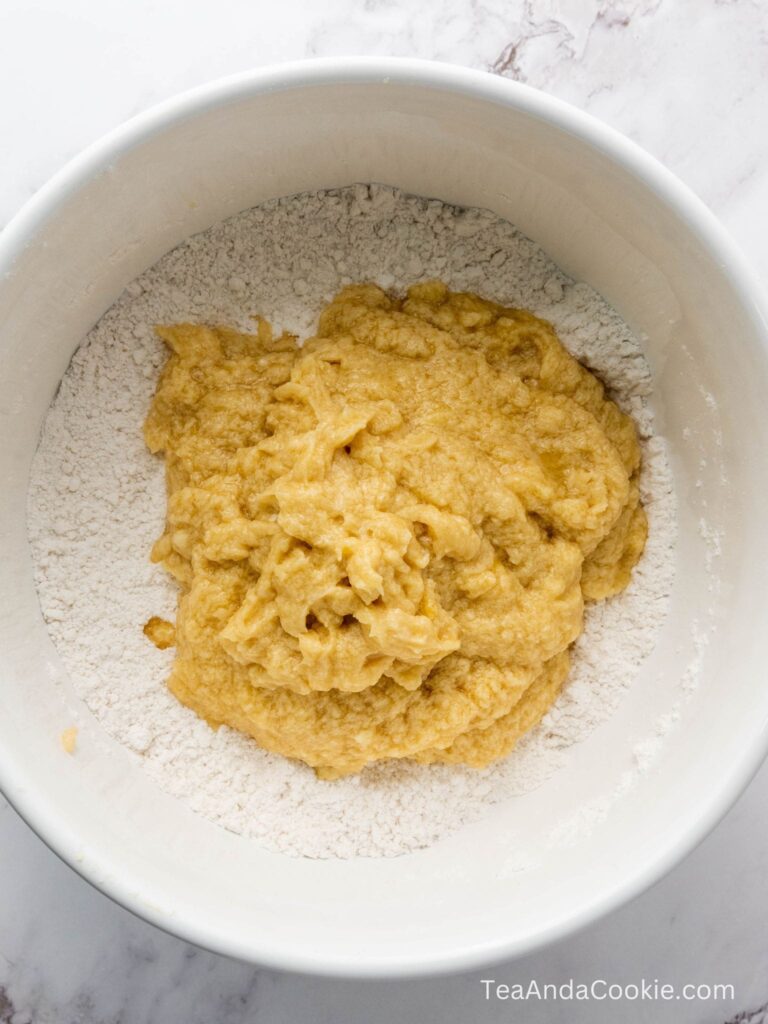 A mixing bowl with flour at the bottom and a mound of wet, yellowish dough on top, ready to be mixed together.
