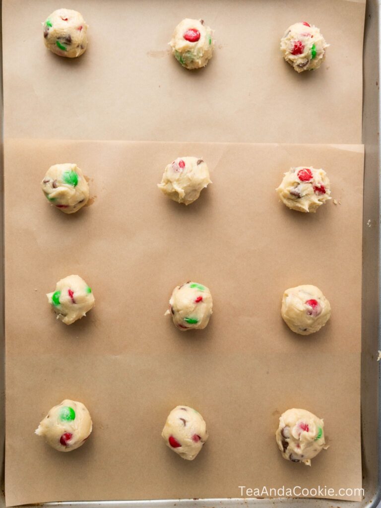 Cookie dough balls with red and green candies arranged in rows on a parchment-lined baking sheet, ready to be baked.