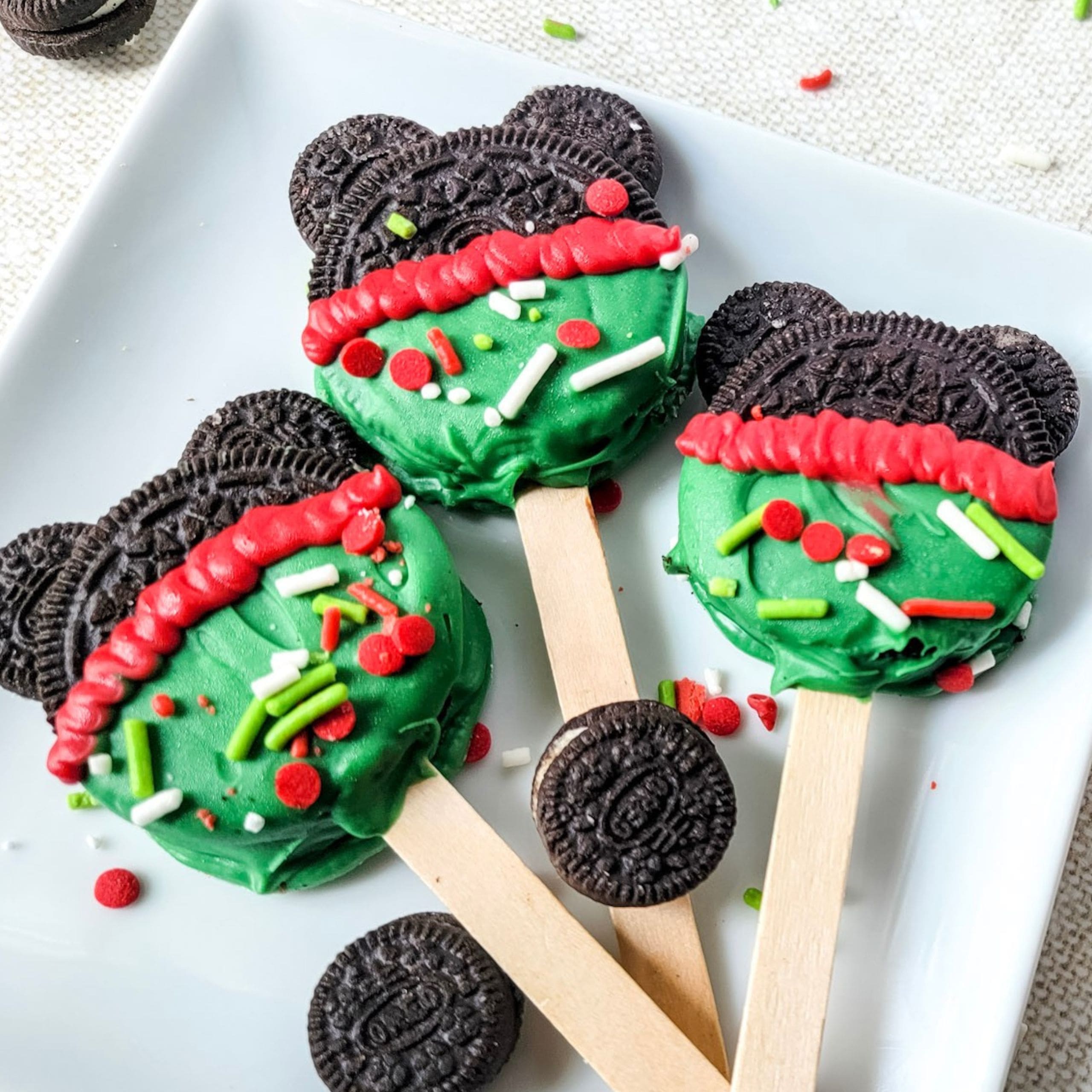 Mickey Christmas Oreos. Three Oreo cookie pops decorated with green and red icing and festive sprinkles are displayed on a white plate, with popsicle sticks as handles.