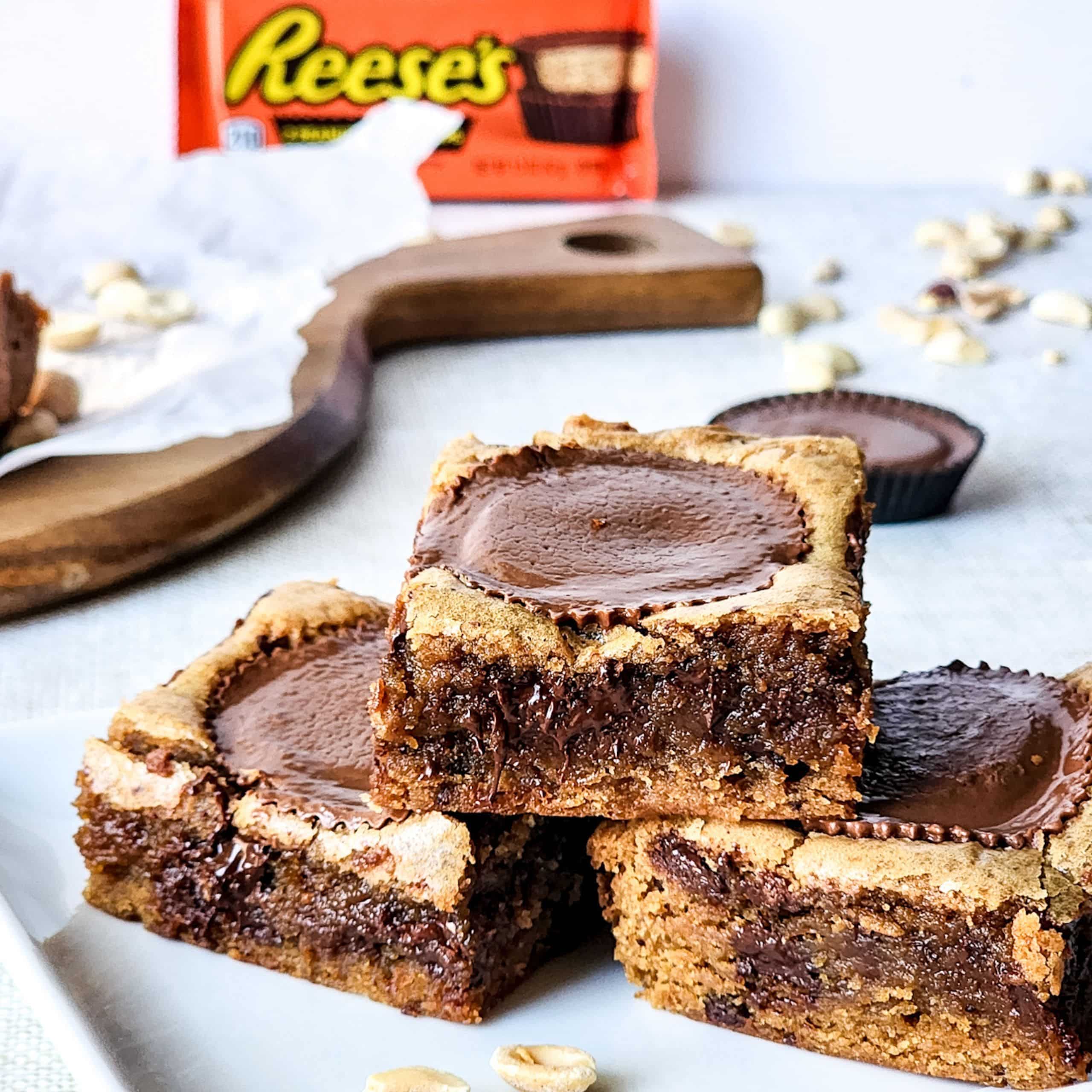 Three chocolate peanut butter cookie bars with Reeses cups in the center, stacked on a white plate, with a Reeses package and peanuts in the background.
