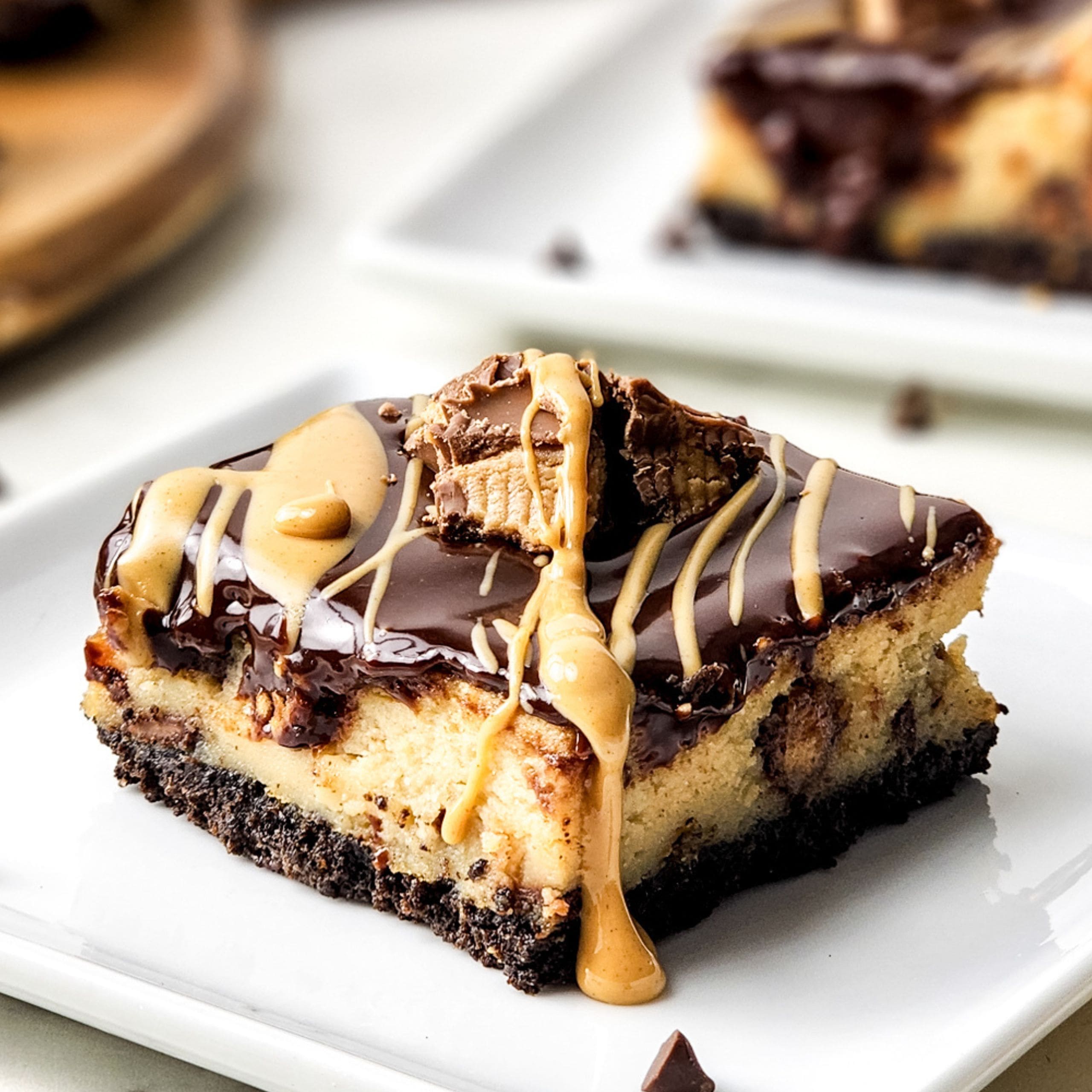 A square piece of cheesecake with a chocolate cookie crust, topped with chocolate ganache, peanut butter drizzle, and a chunk of peanut butter cup, served on a white plate.