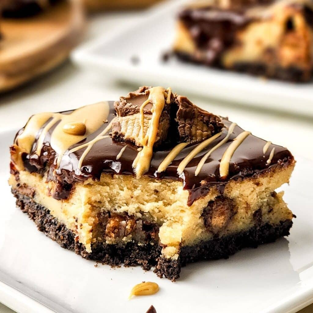 Peanut butter cheesecake bars. A slice of cheesecake with a chocolate cookie crust, topped with chocolate ganache, peanut butter drizzle, and pieces of peanut butter cup candy on a white plate.