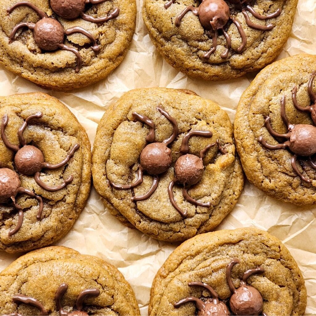 Chocolate chip cookies decorated with chocolate balls and icing to resemble spiders, arranged on crumpled parchment paper.