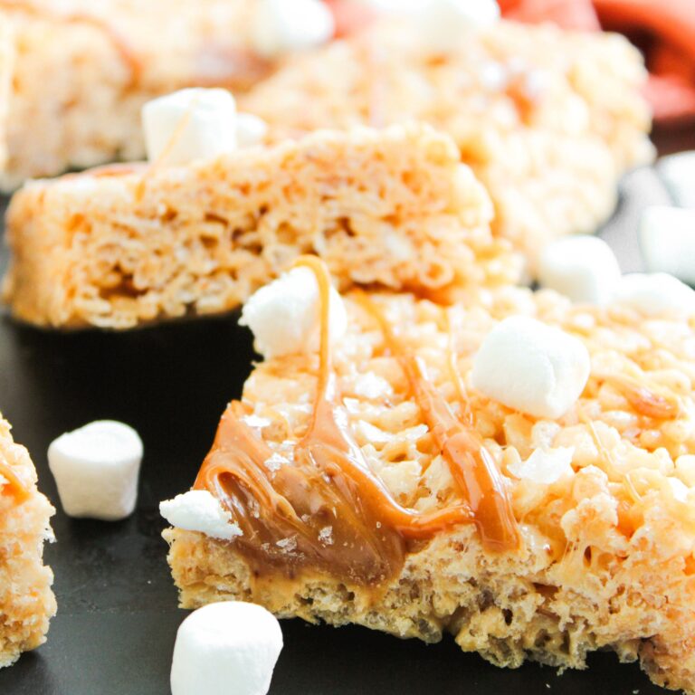 Close-up of rice crispy treats drizzled with caramel and topped with mini marshmallows, with more marshmallows scattered around.