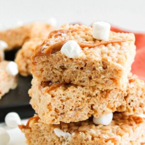 Three rice krispie treats stacked on top of each other, drizzled with caramel and topped with small marshmallows.
