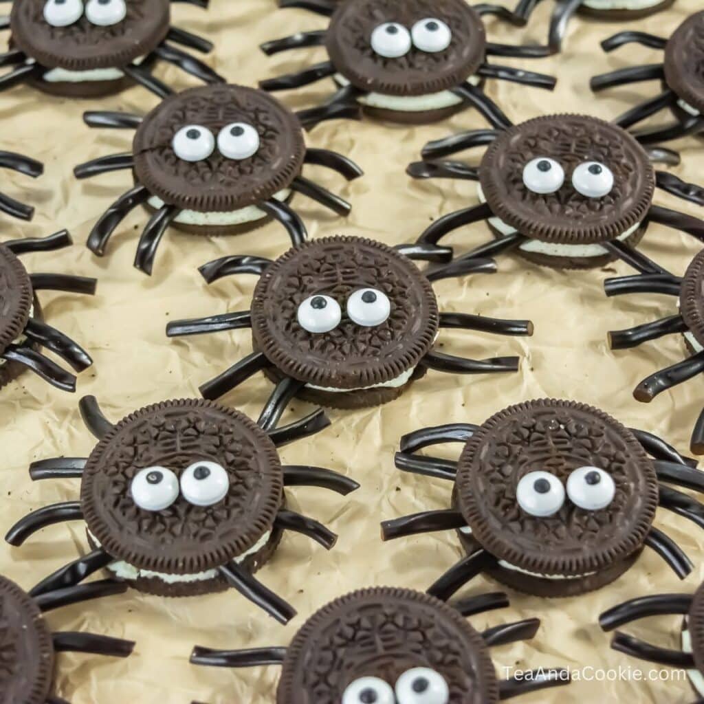 Chocolate sandwich cookies decorated with candy eyes and black licorice legs to resemble spiders, arranged on a crinkled parchment surface.