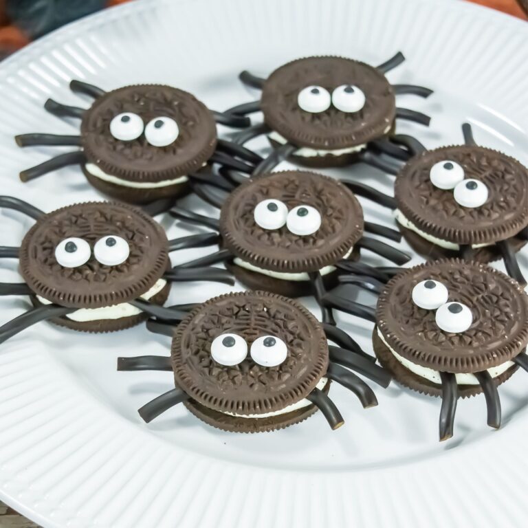 A plate of cookies decorated to look like spiders, with candy eyes and black licorice pieces for legs, arranged on a white plate.