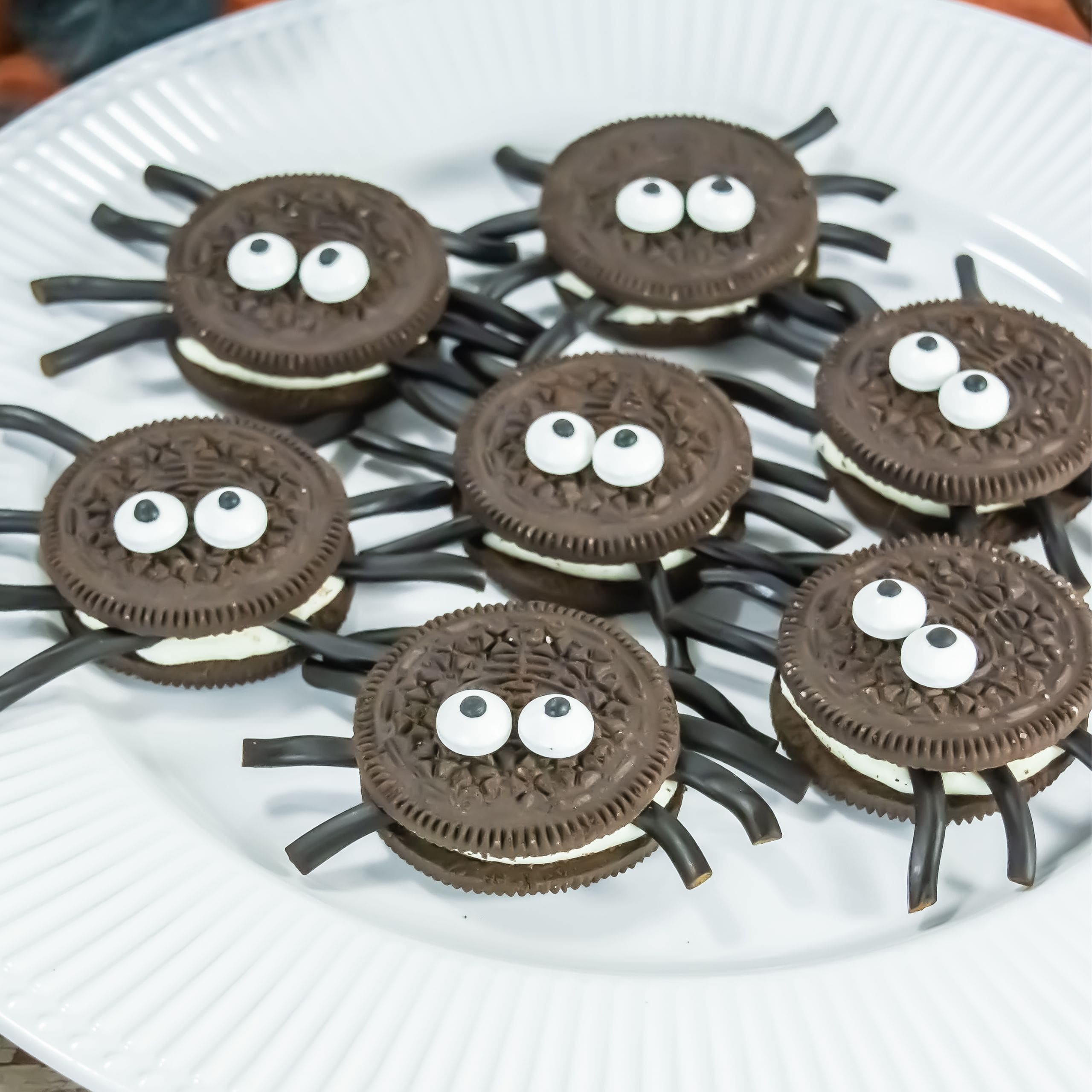 A plate of cookies decorated to look like spiders, with candy eyes and black licorice pieces for legs, arranged on a white plate.