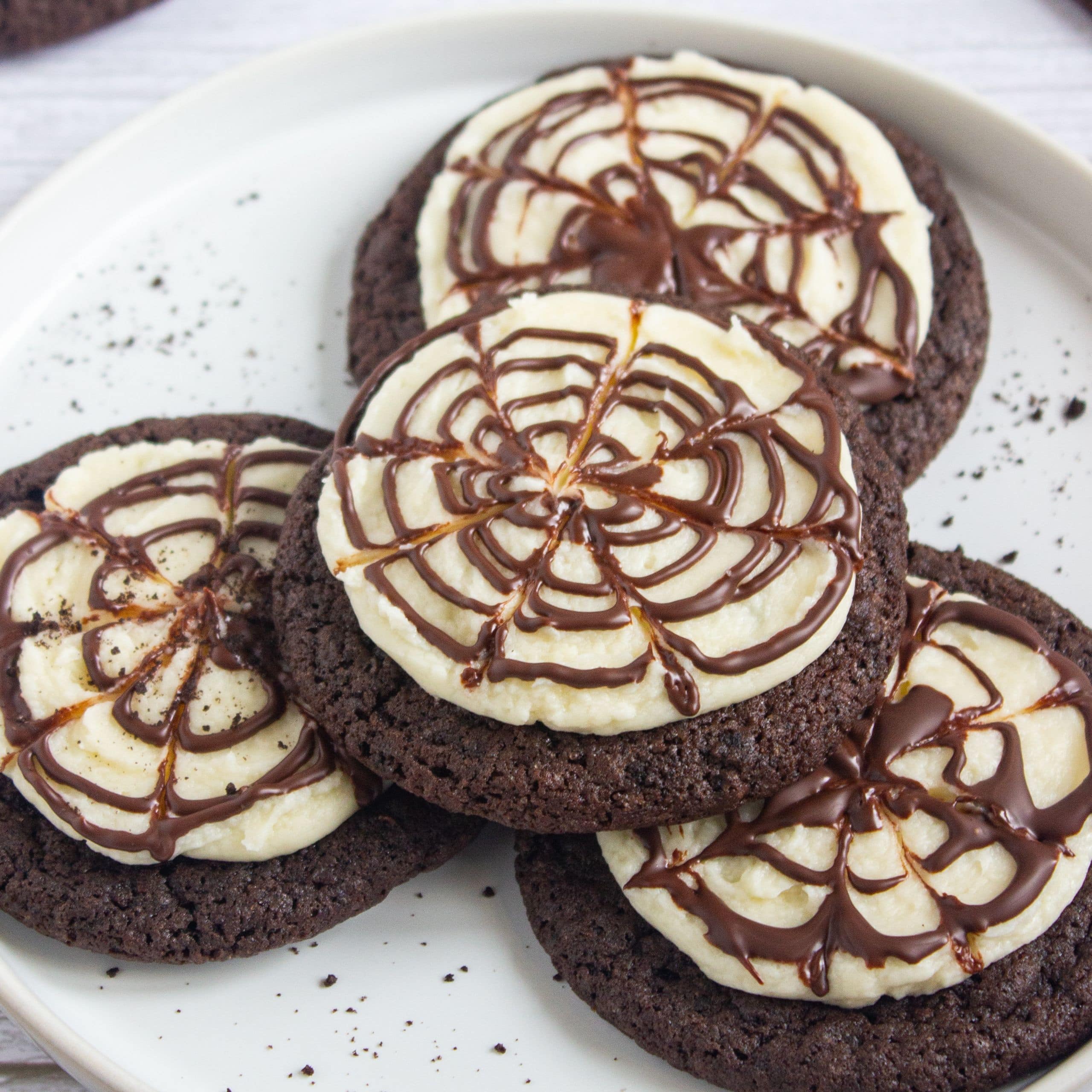 Four chocolate cookies topped with white icing and a chocolate spiderweb design are arranged on a white plate.