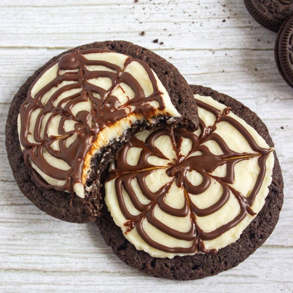 Spiderweb Cookies. Two chocolate cookies with white icing and chocolate spiderweb designs; one cookie has a bite taken out of it.