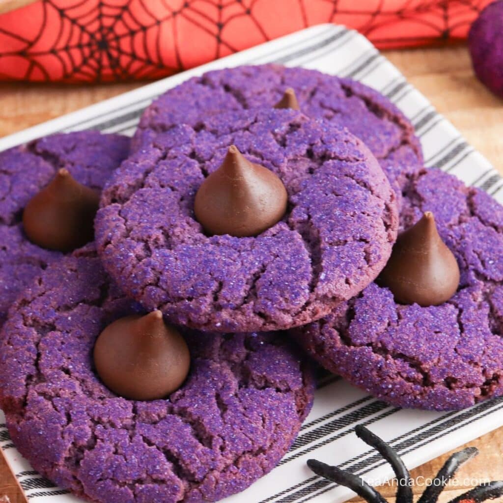 A plate of purple sugar cookies, each topped with a chocolate drop, is displayed on a striped tray with a Halloween-themed napkin in the background.