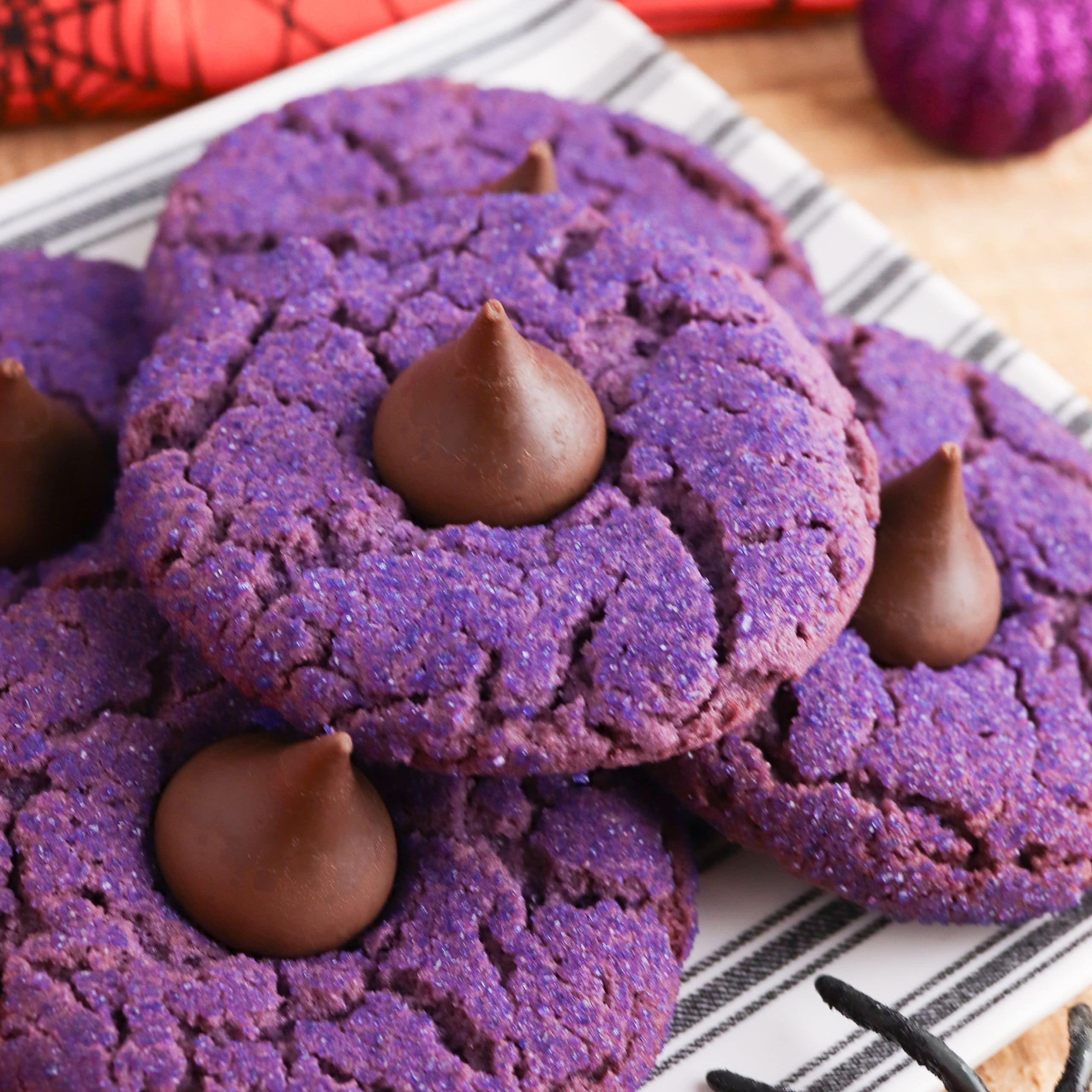 A stack of purple sugar cookies topped with chocolate kisses on a striped plate, with Halloween decorations in the background.