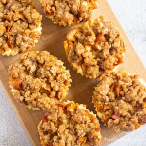 Six apple crumble muffins with a crumbly topping are arranged on a wooden board.