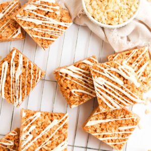 Rice crispy treats with white chocolate drizzle are arranged on a cooling rack, with a bowl of crispy rice cereal in the background.