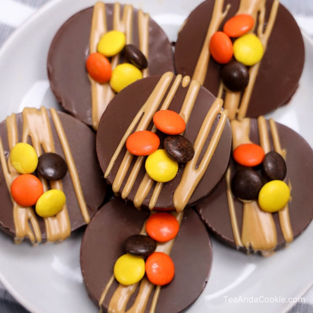 Chocolate covered Oreos with Peanut Butter. Five round chocolate discs topped with a drizzle of peanut butter and decorated with orange, yellow, and brown candy-coated chocolates, arranged on a white plate.