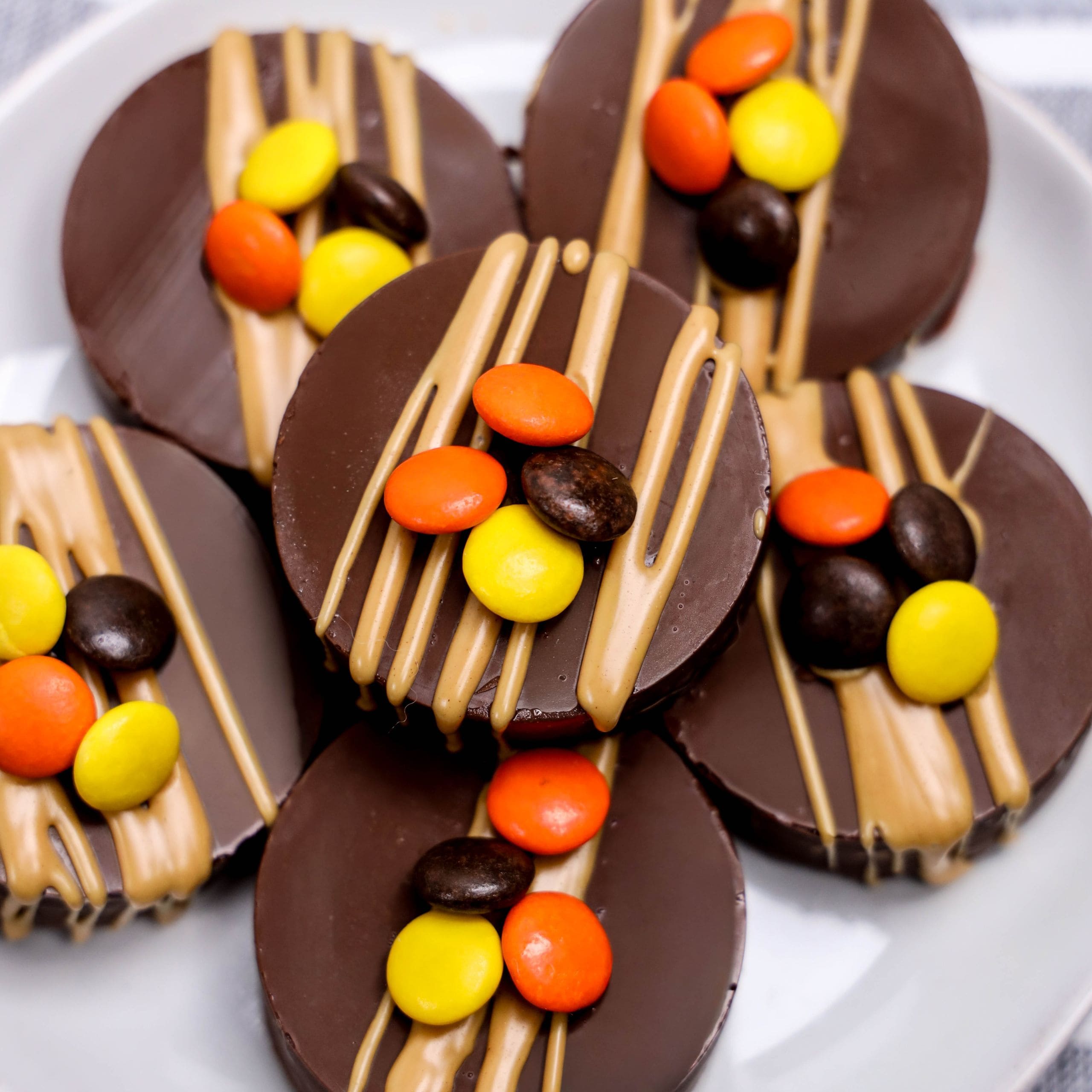 Five round chocolate discs topped with tan-colored drizzle and orange, yellow, and brown candy-coated chocolates, arranged on a white plate.