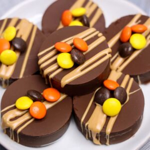 Five round chocolate-covered treats are topped with tan drizzle and colorful orange, yellow, and brown candy-coated chocolates, arranged on a white plate.
