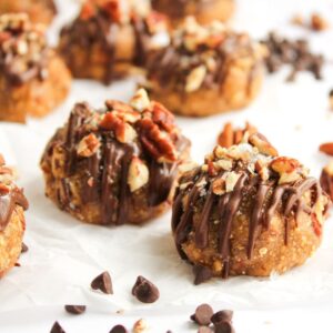 Round cookies topped with chopped nuts and drizzled with chocolate are arranged on parchment paper with chocolate chips scattered nearby.
