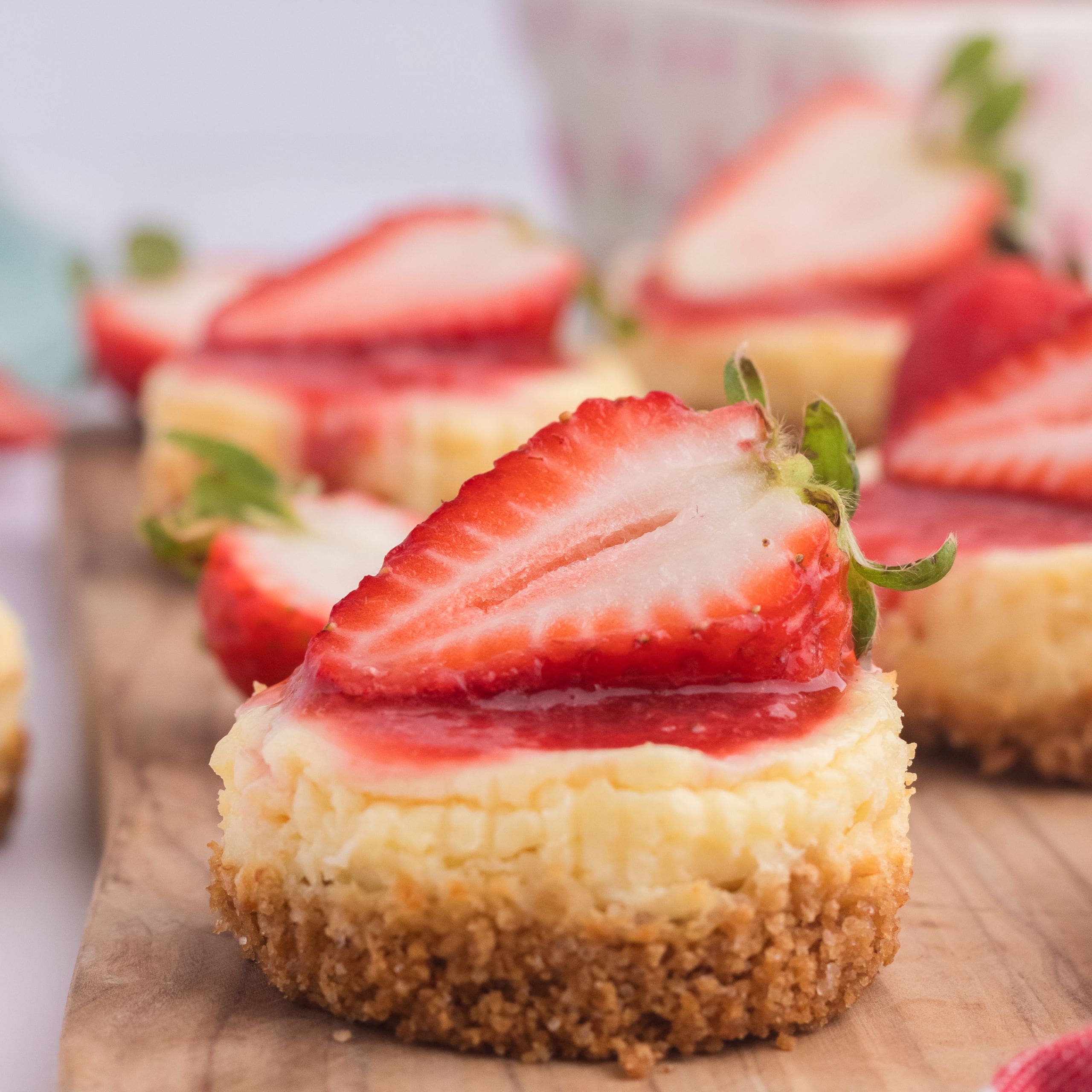 A mini cheesecake with a graham cracker crust, topped with a fresh strawberry slice, sits on a wooden board with more cheesecakes in the background.
