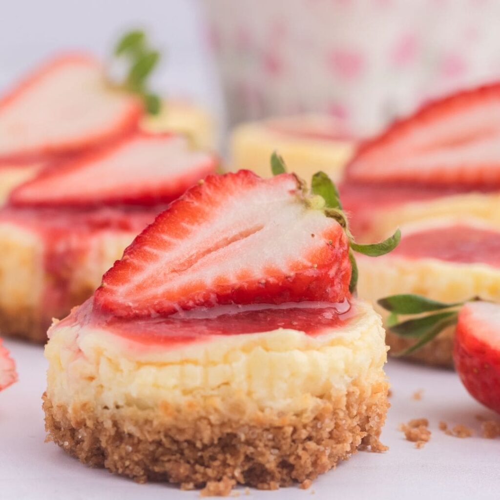 Miniature strawberry cheesecakes. Mini cheesecakes topped with strawberry slices and sauce, each with a crumbly crust, arranged on a white surface.