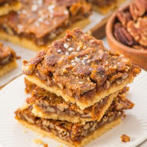 Three pecan pie bars stacked on a white plate, with a bowl of pecans and more bars in the background.