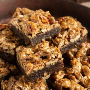 A close-up of stacked chocolate brownies topped with a layer of pecans and a caramelized glaze on a brown plate.