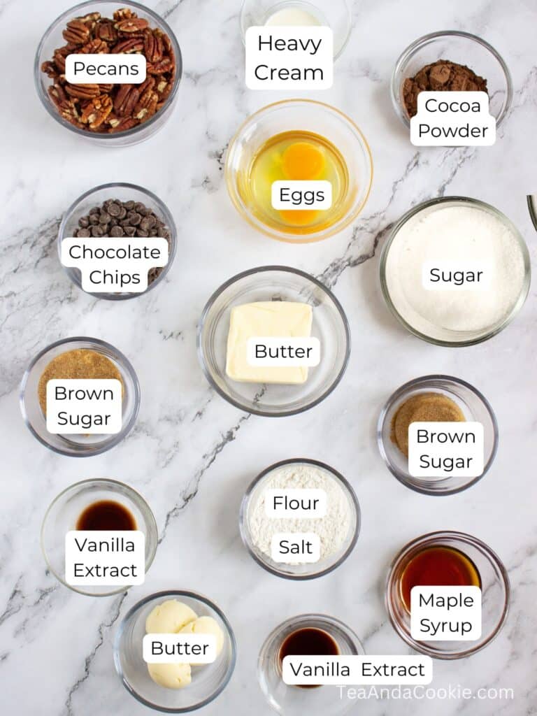 Various labeled baking ingredients, including pecans, heavy cream, eggs, cocoa powder, chocolate chips, sugars, butter, vanilla extract, flour, salt, and maple syrup, arranged on a marble surface.
