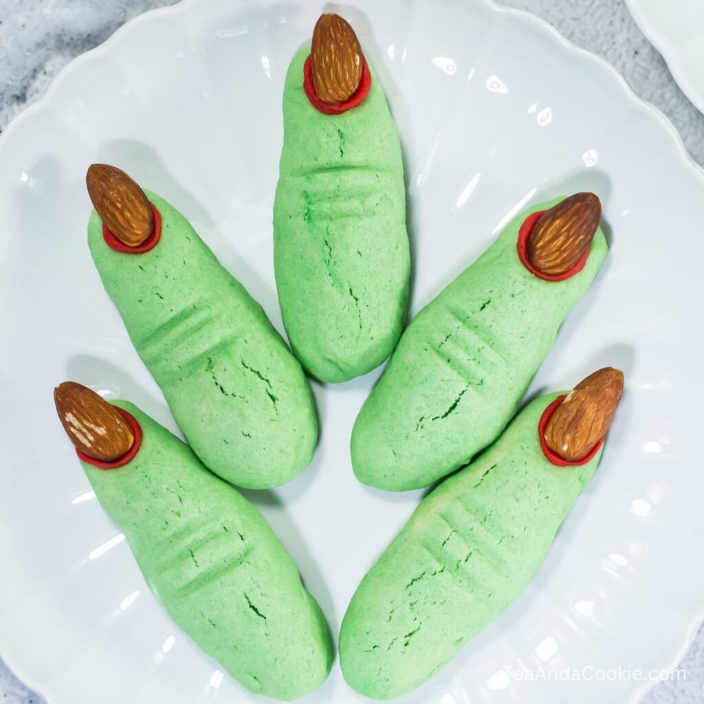 Shortbread Witch Finger Cookies Recipe. Five green cookies shaped like fingers with almond nails and red detailing are arranged on a white plate.
