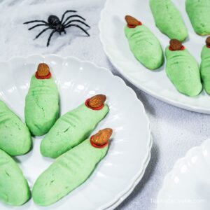 Two white plates hold green cookies shaped like fingers with almond nails and red icing, alongside a black plastic spider decoration.