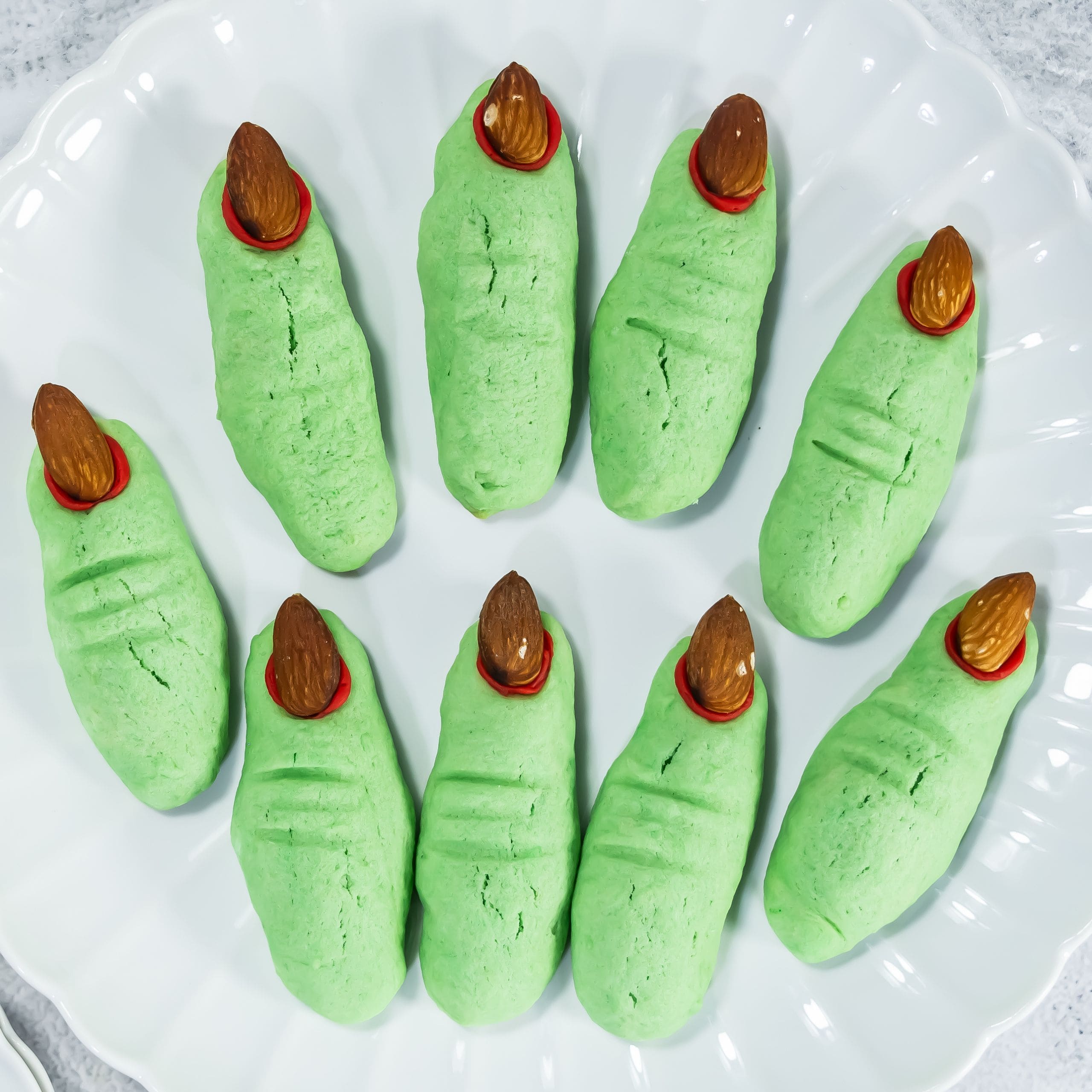 Green cookies shaped like fingers with almond “nails” and red icing detail, arranged in a circular pattern on a white plate.